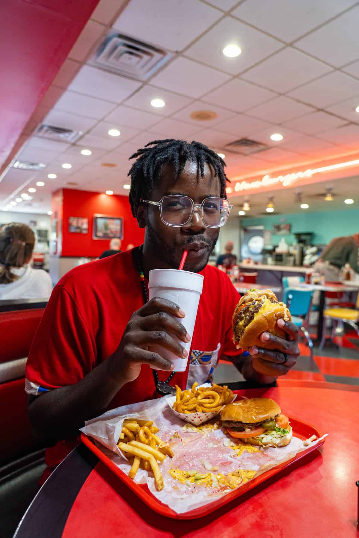 a man in a red shirt drinking a drink and eating a burger