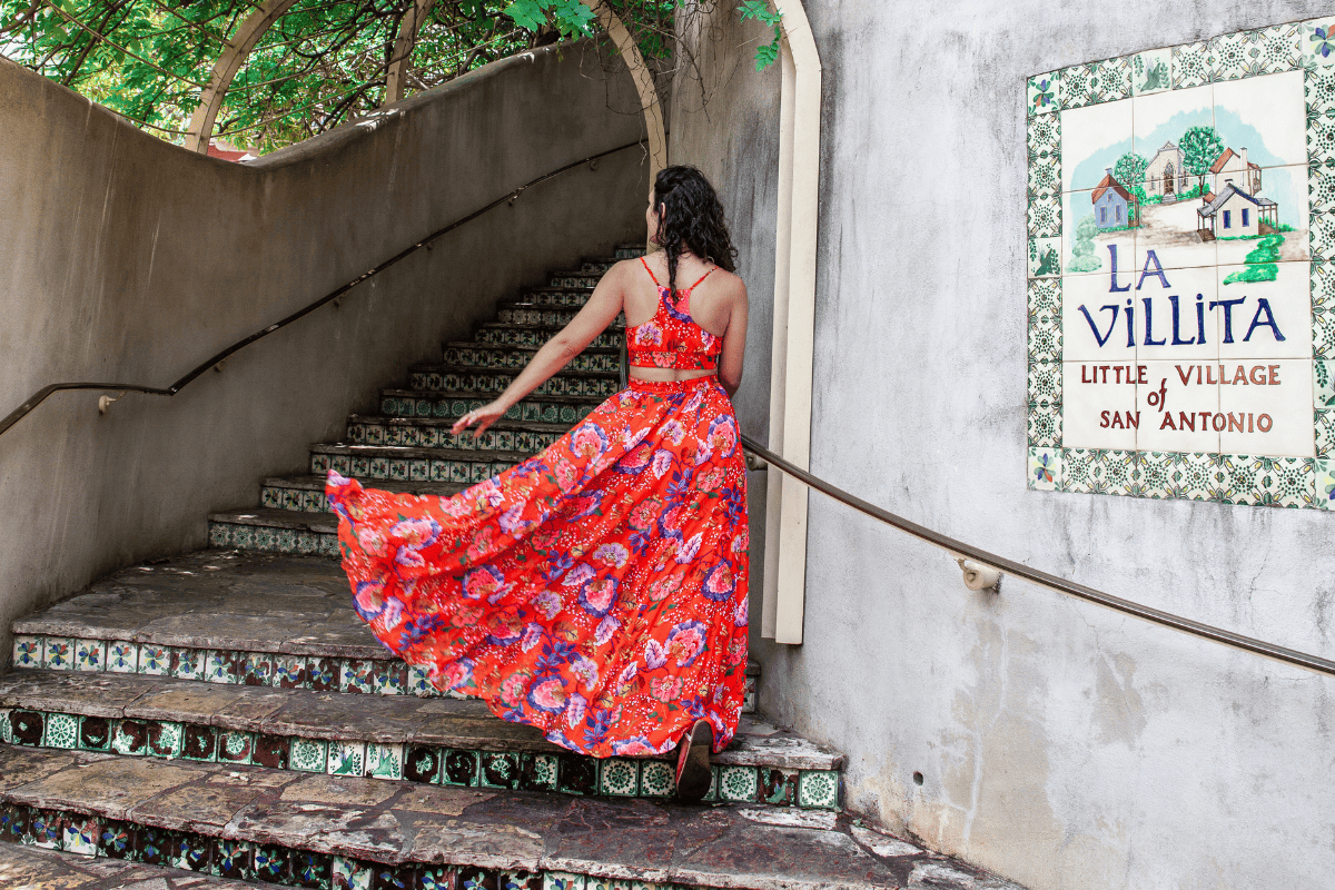 Woman in red floral dress ascending tiled stairs by "La Villita" sign.