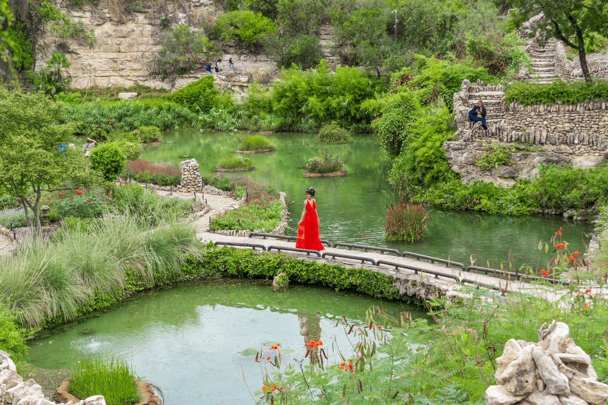 Woman wearing red dress crosses bridge over pond.