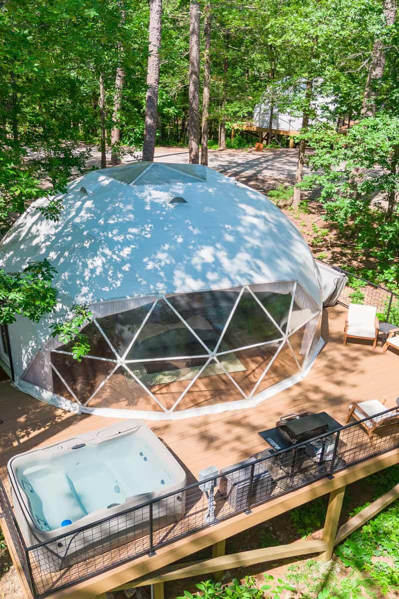 Geodesic dome house on a wooden deck with a hot tub and grill in a forest setting.