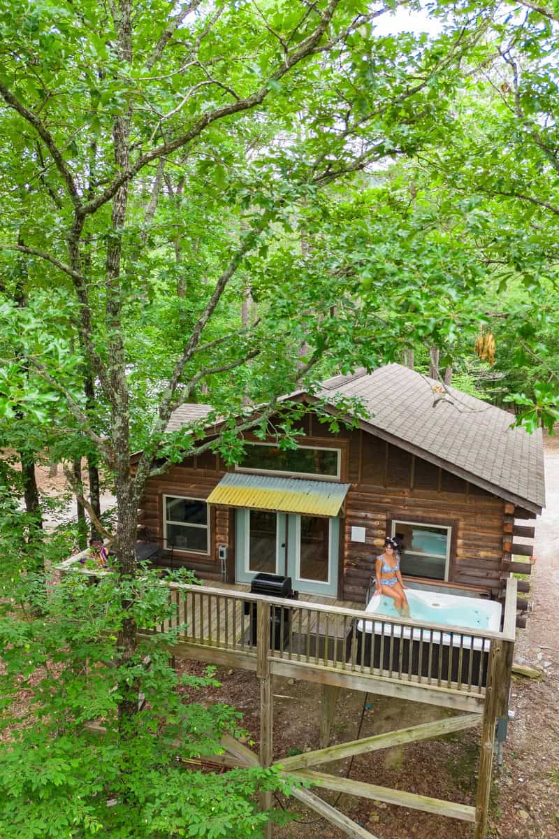 Cabin in woods with elevated deck and person in swimsuit standing by a hot tub.