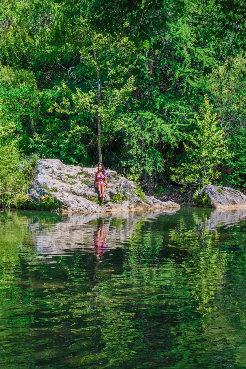 Person sitting on a rocky outcrop by a tranquil, tree-lined river.