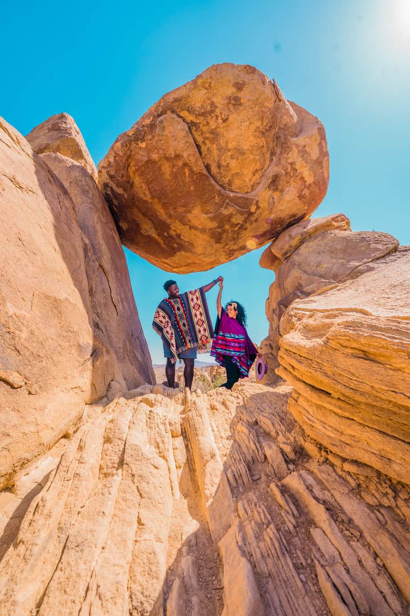 Two people with patterned garments standing under a balanced rock formation 