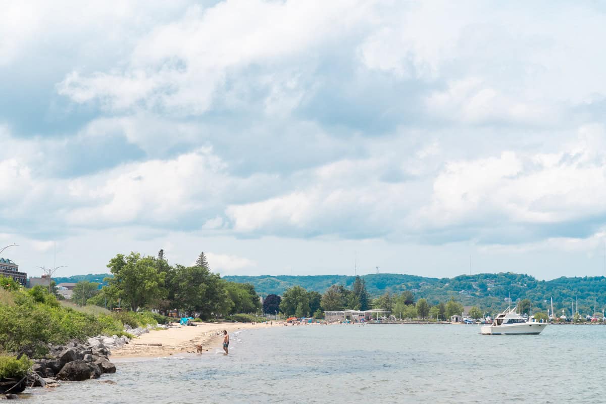 Sandy beach with people by the water, a boat nearby, against a backdrop of hills and clouds.