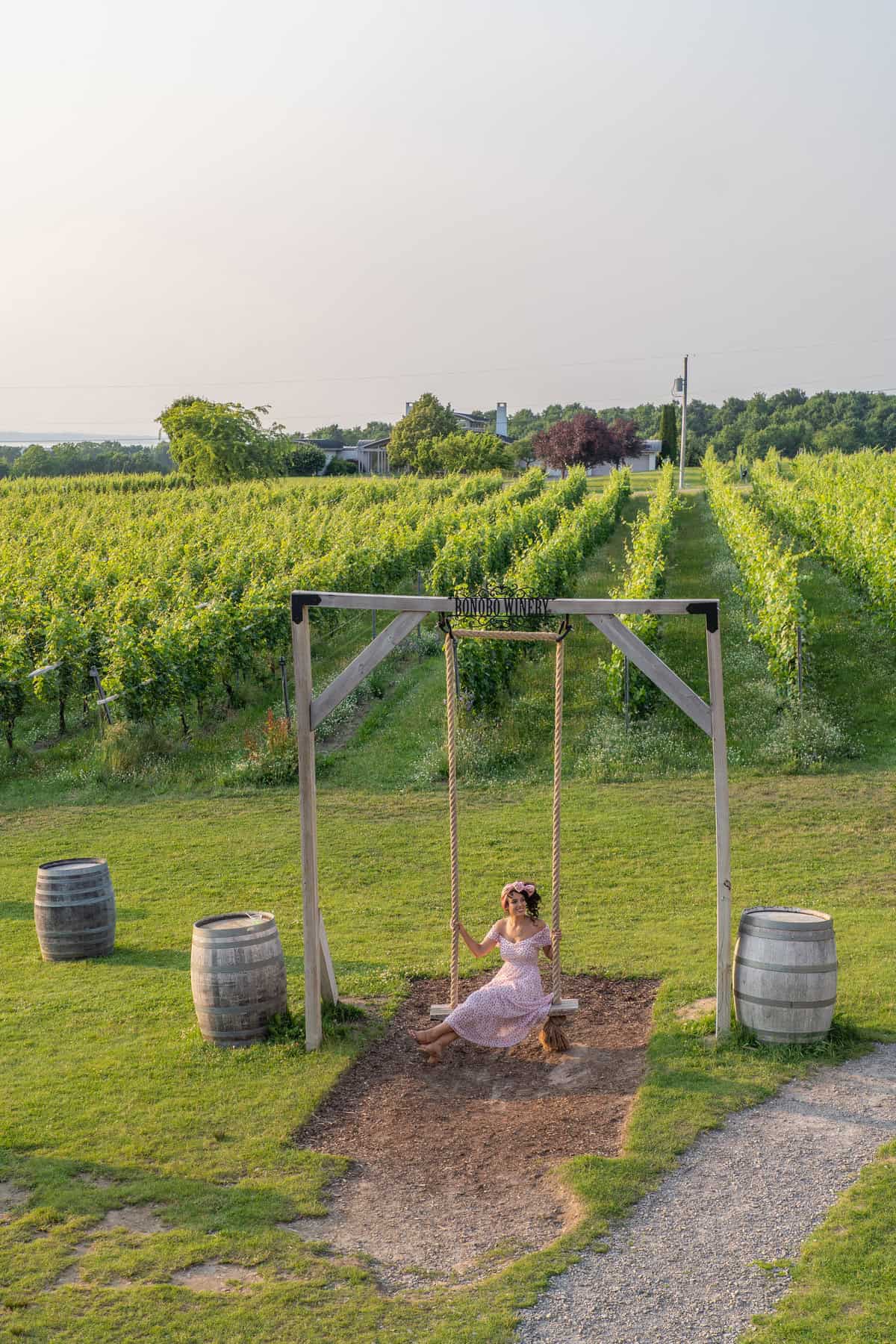 Person in a dress on a swing at a vineyard with wine barrels and greenery.