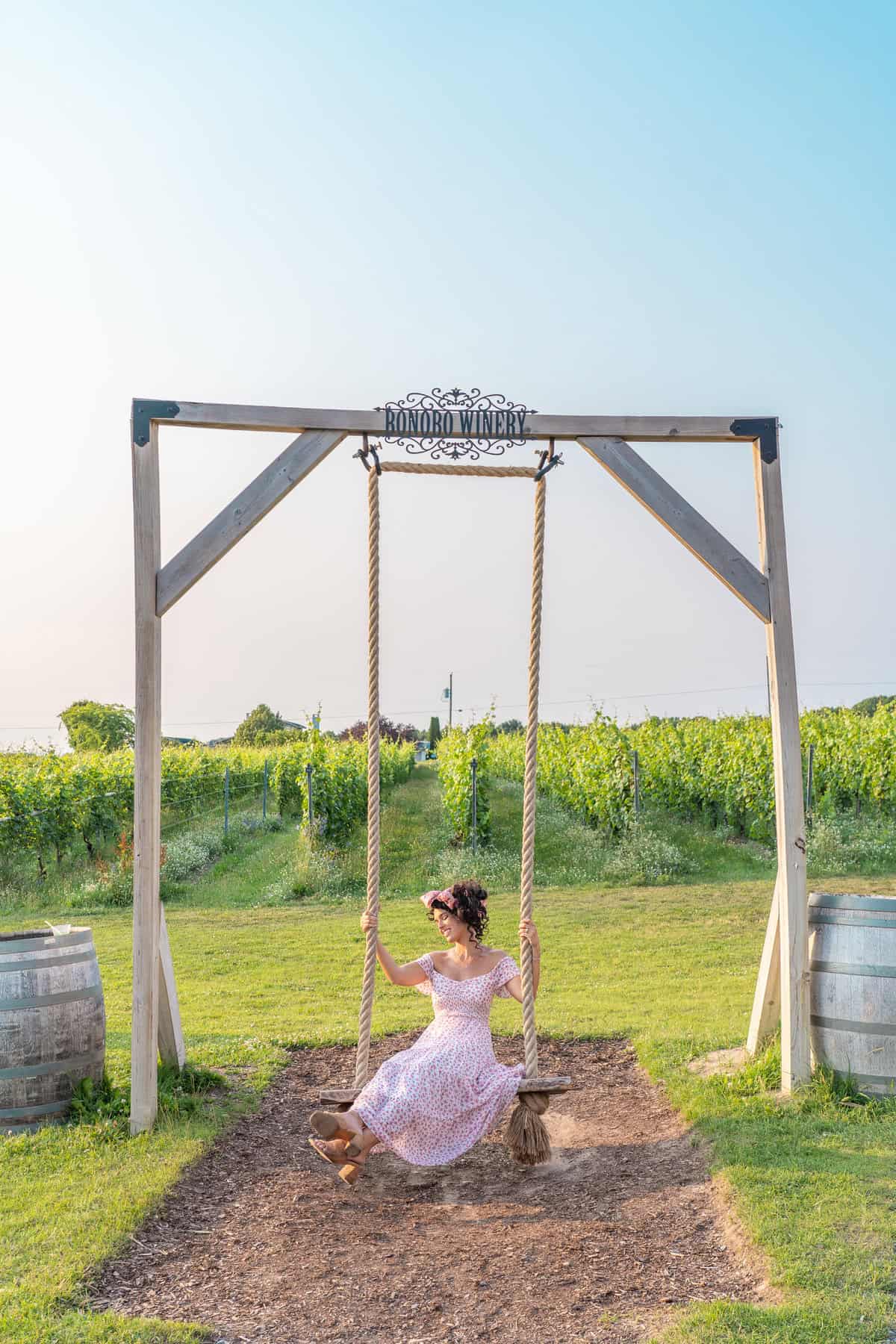 Woman in a dress on a wooden swing at a vineyard with clear skies.