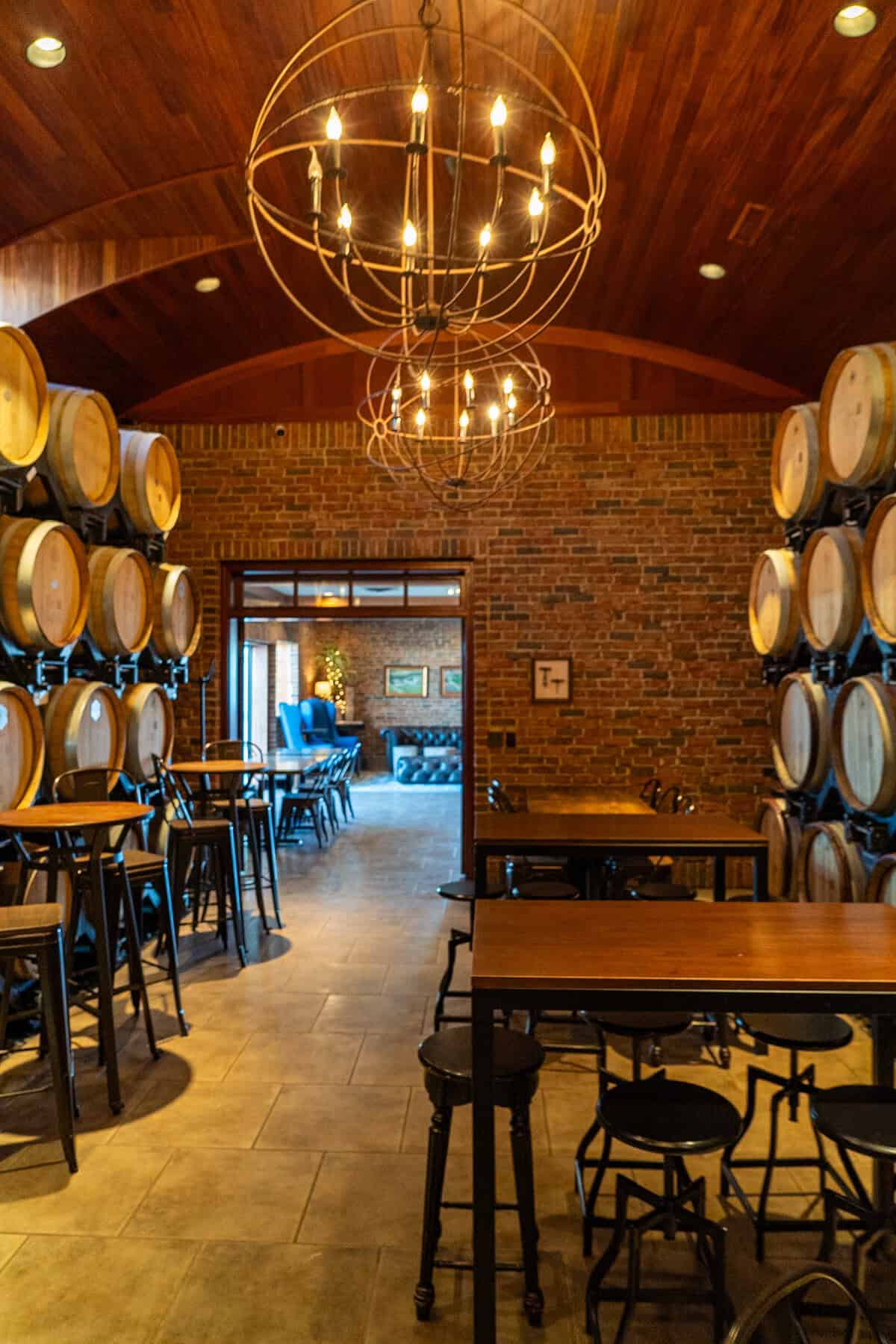 A cozy winery tasting room with wooden barrels, brick walls, and circular chandeliers.