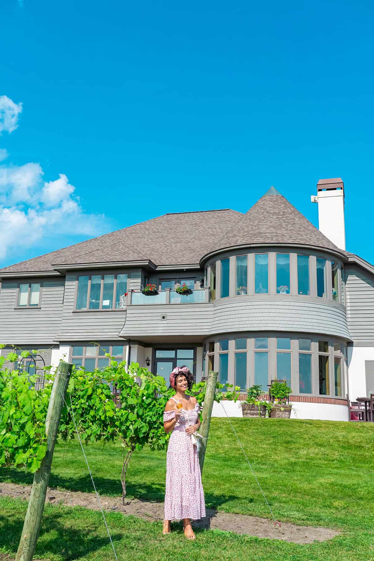 Person in a pink dress holding a book near vine rows in front of a large house 