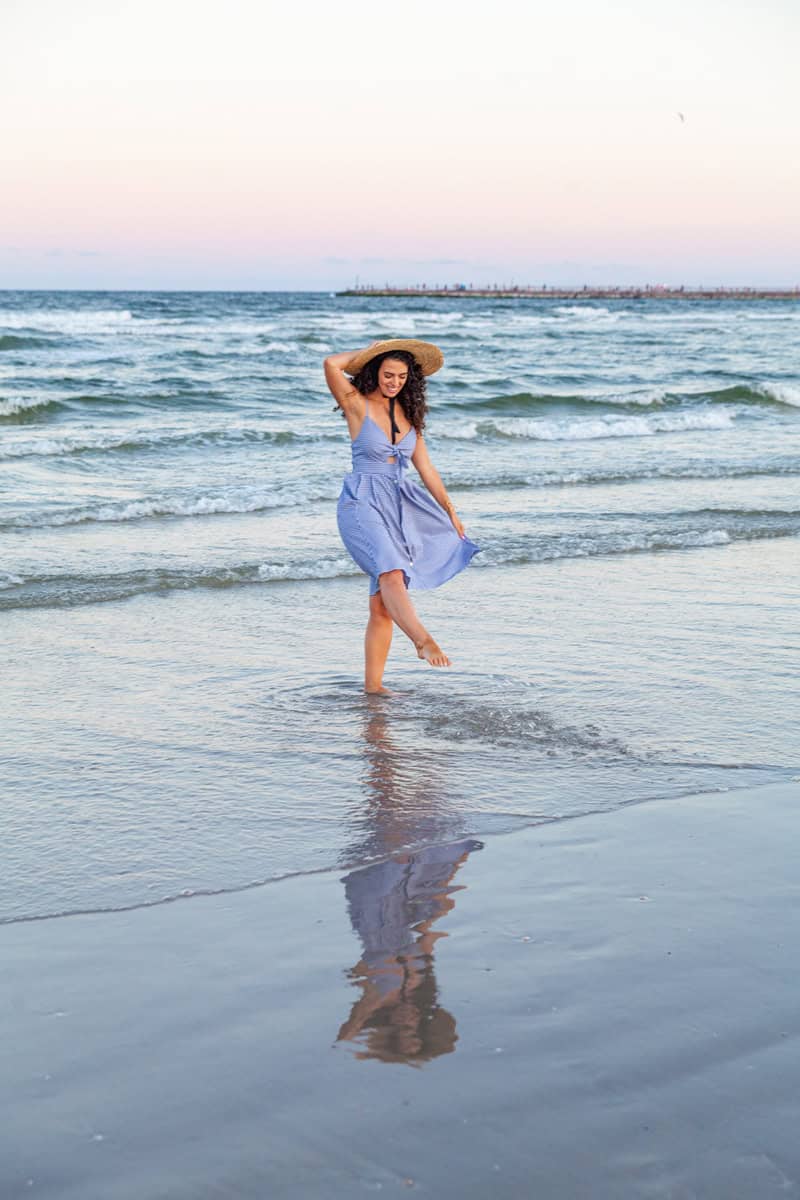 Person in a blue dress and straw hat standing on the beach at sunset.