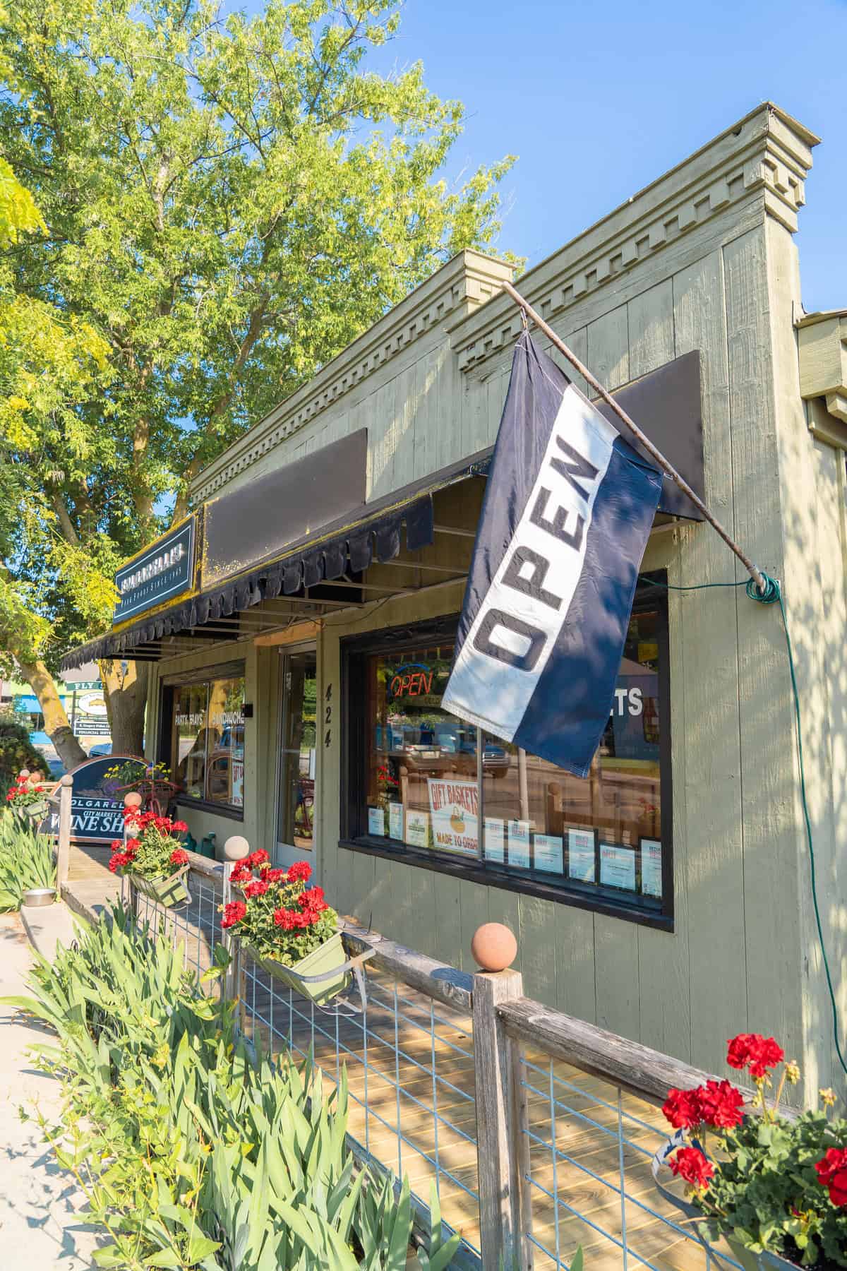 Storefront with 'OPEN' flag and red flowers under a clear sky.