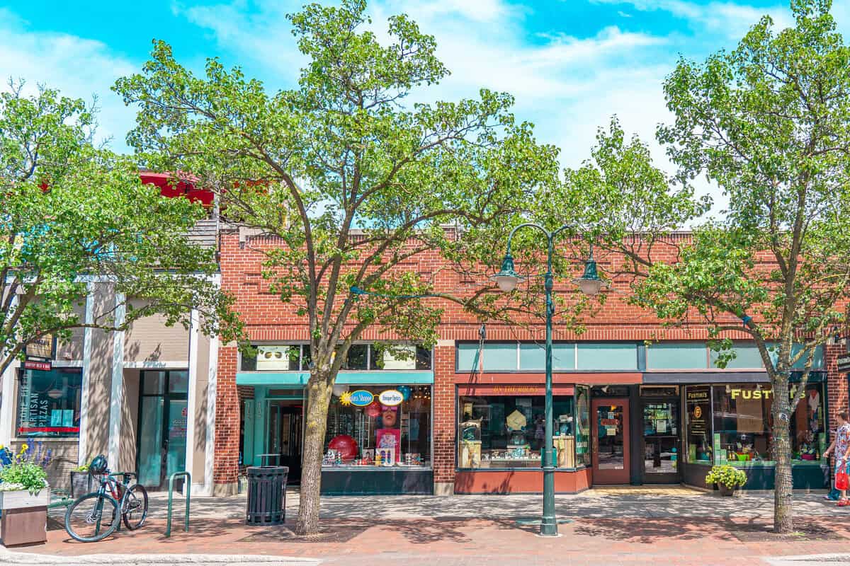 A sunny street view of a brick building with shops, green trees, and a blue sky.