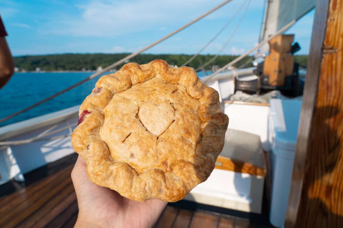 A hand holding a pie with a heart design onboard a boat with clear skies and water background.