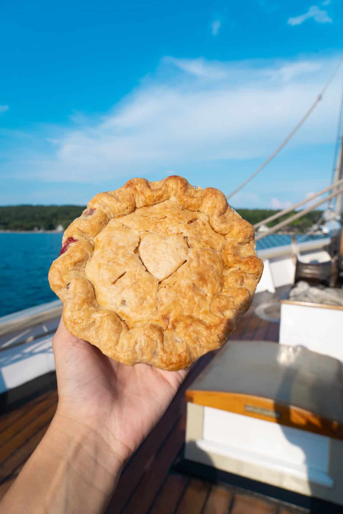 Hand holding a pie with a heart cutout on a boat with blue sky in the background.