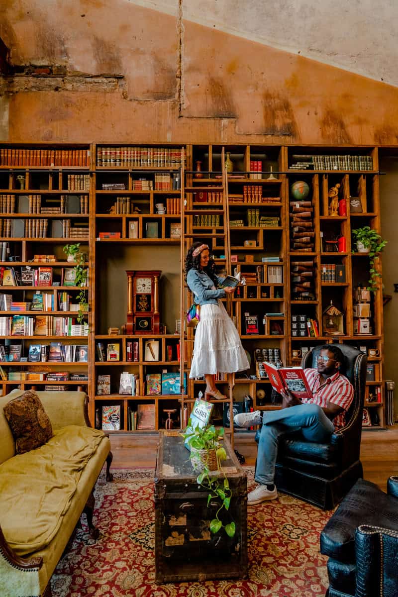 A cozy room with individuals reading by a large bookshelf and vintage furniture.