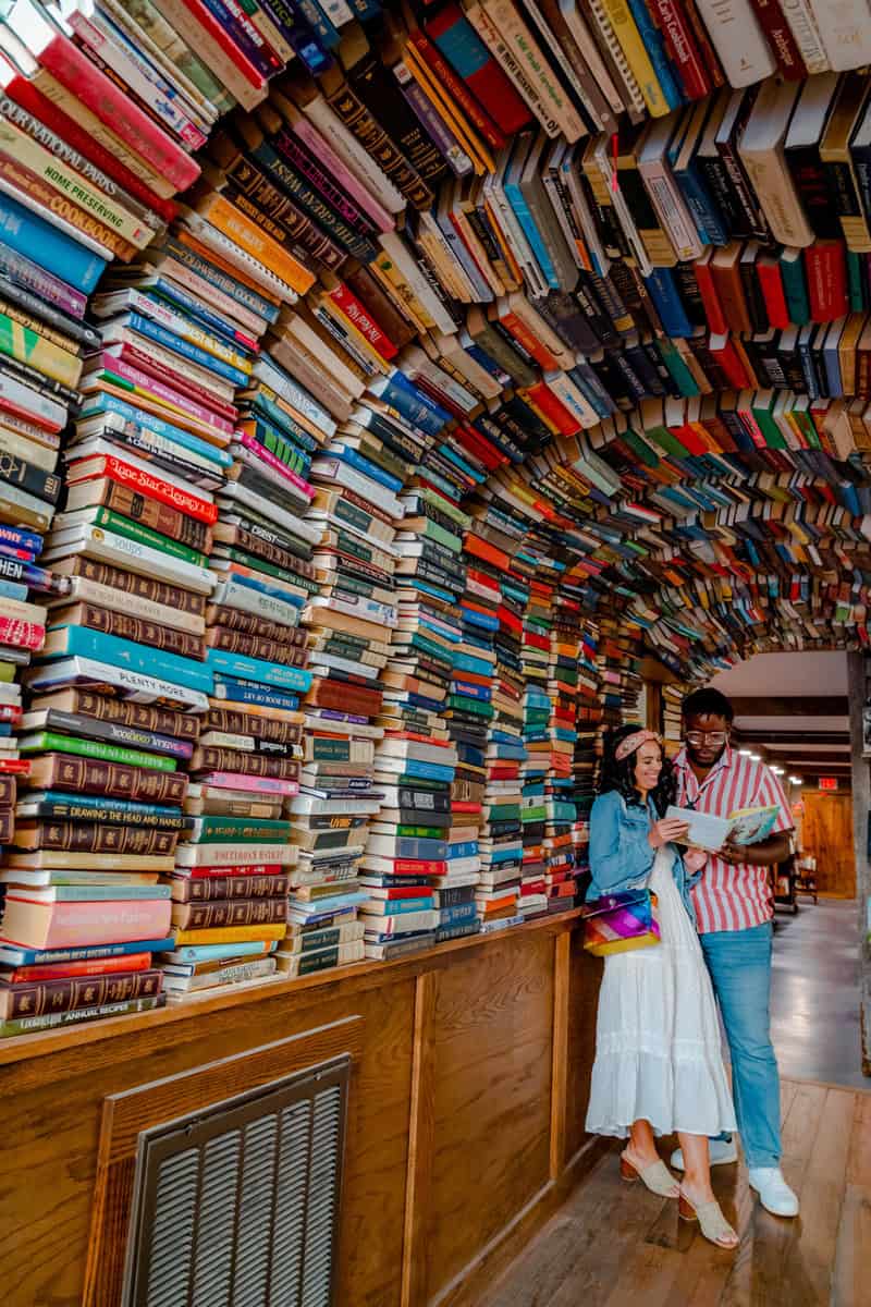 A tunnel of stacked books arching overhead with two individuals standing underneath.