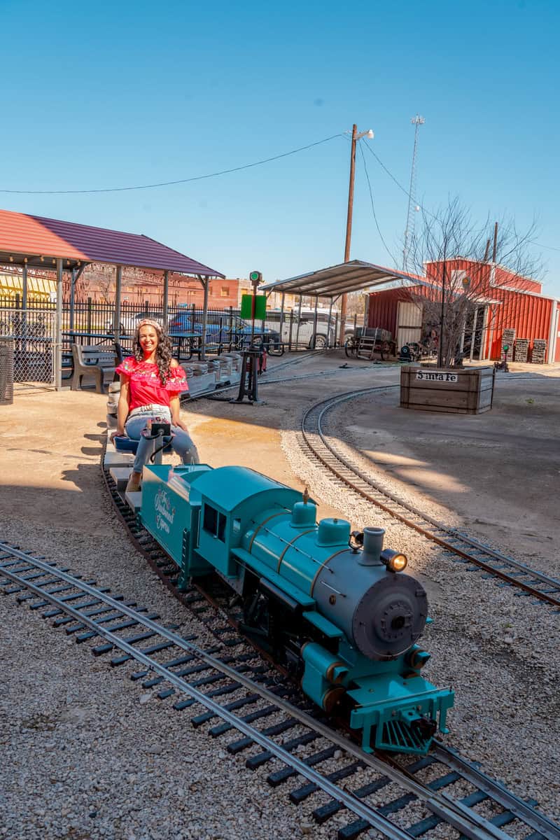 A person rides a miniature blue steam train on a track in a park setting.