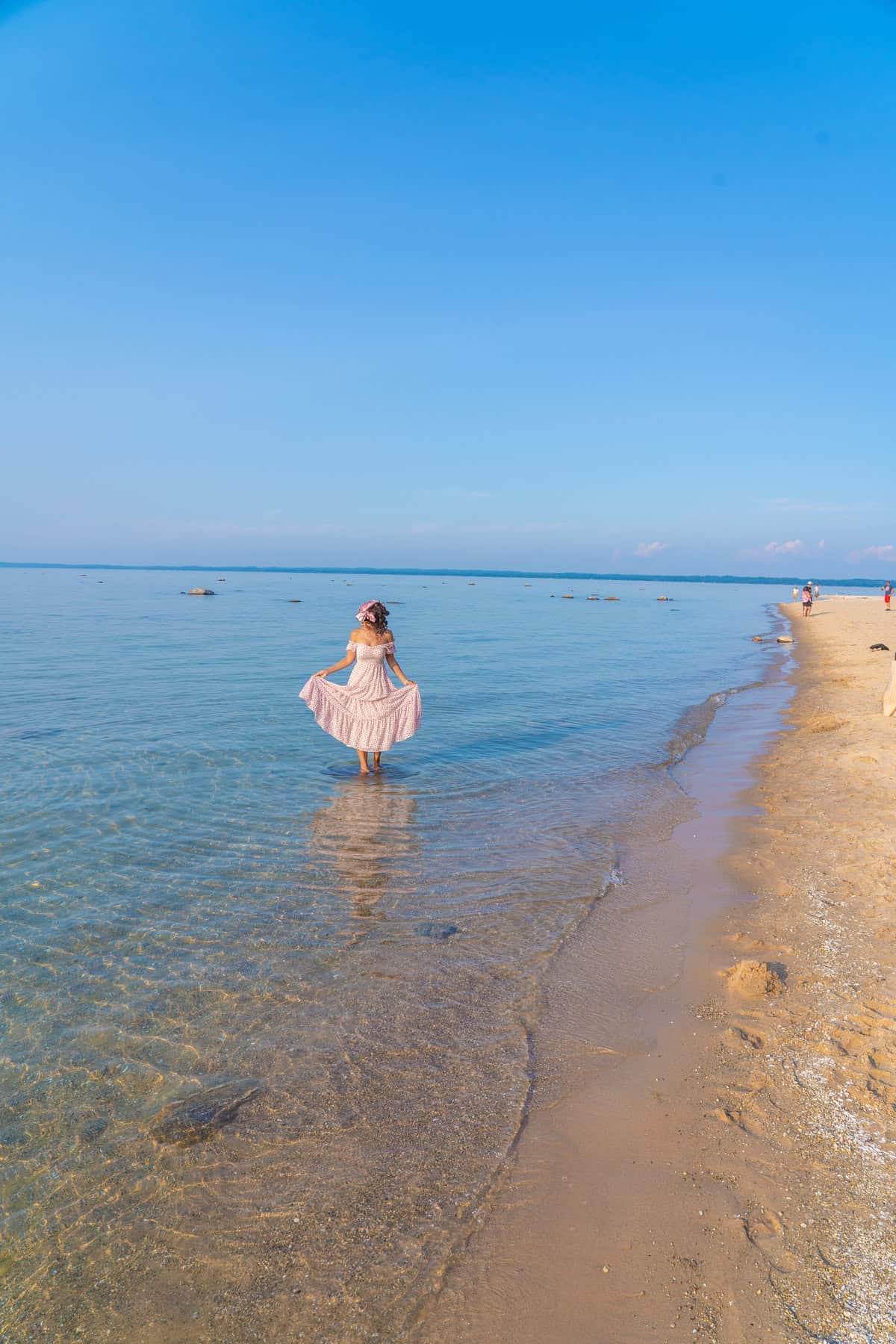 Woman in a pink dress wading in clear, shallow water at a sandy beach with a blue sky.