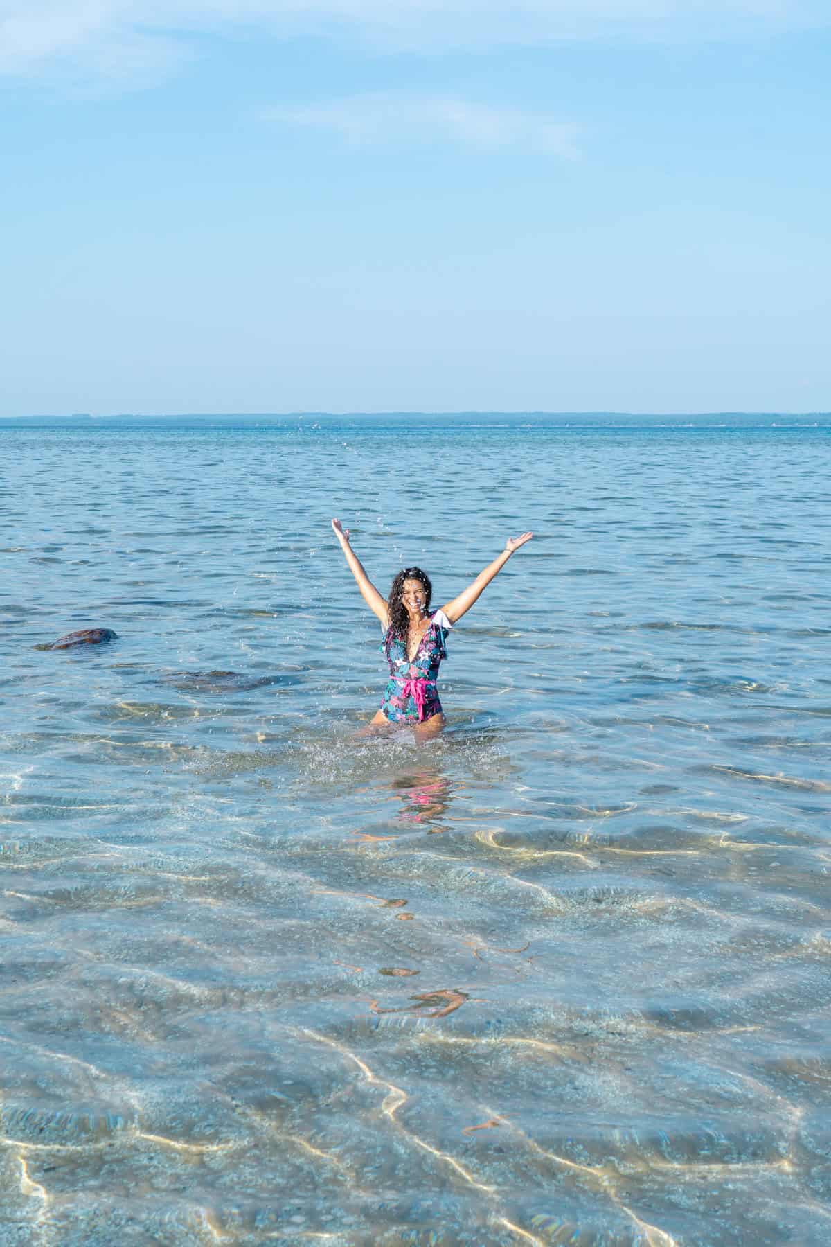 Person with arms raised standing in clear sea water, blue sky above.