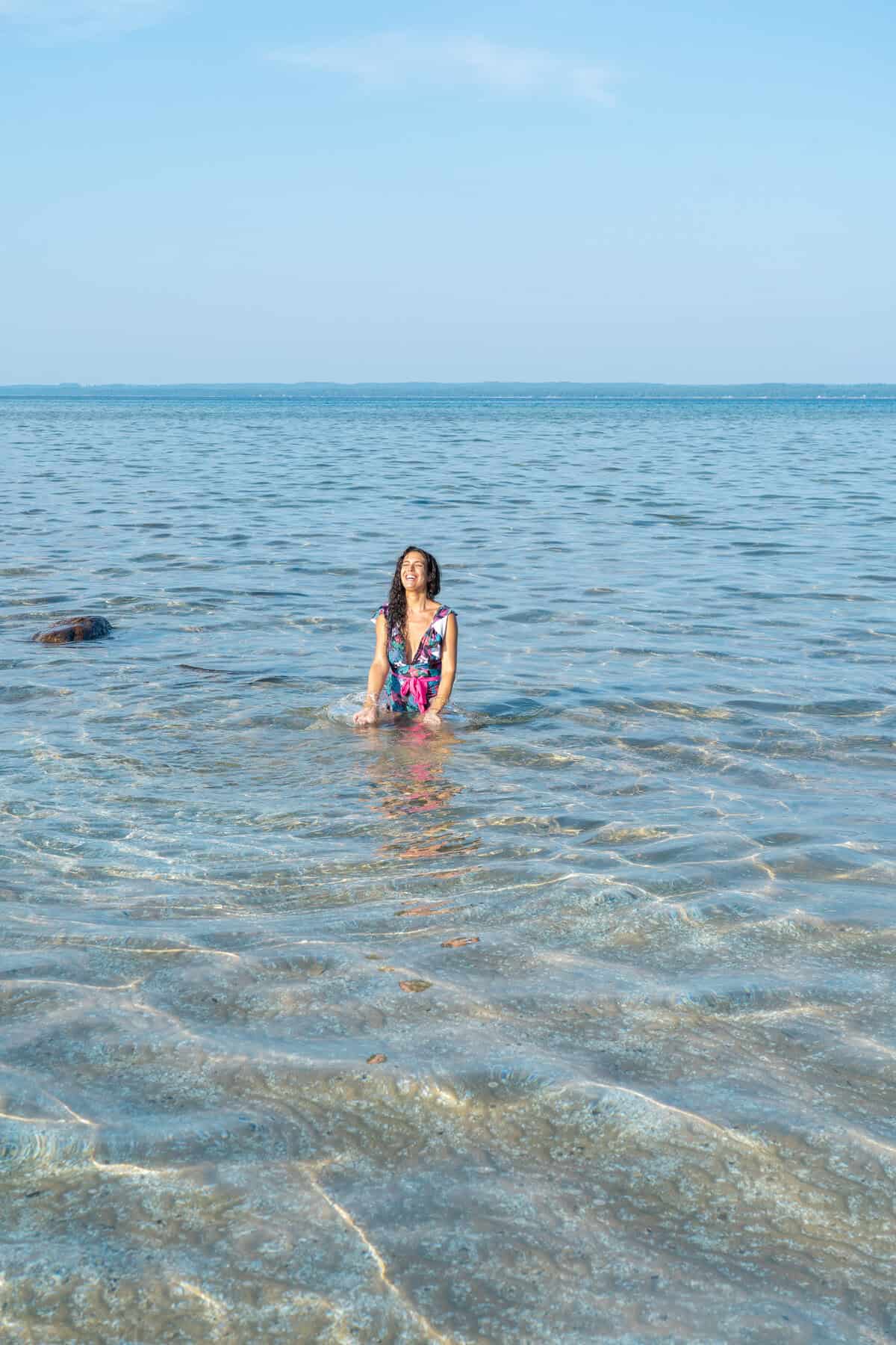 Person in a colorful swimsuit sitting in clear shallow waters of a calm lake.