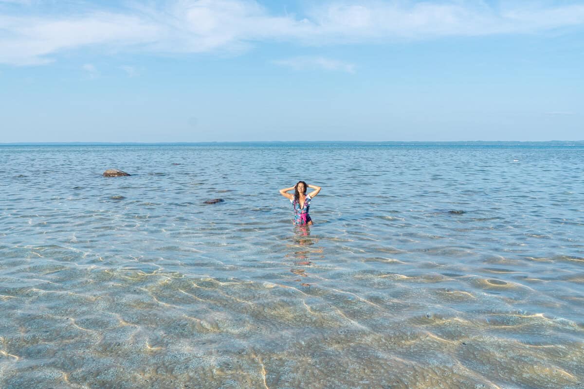 Person standing in clear shallow water, hands on head, with a clear sky above.