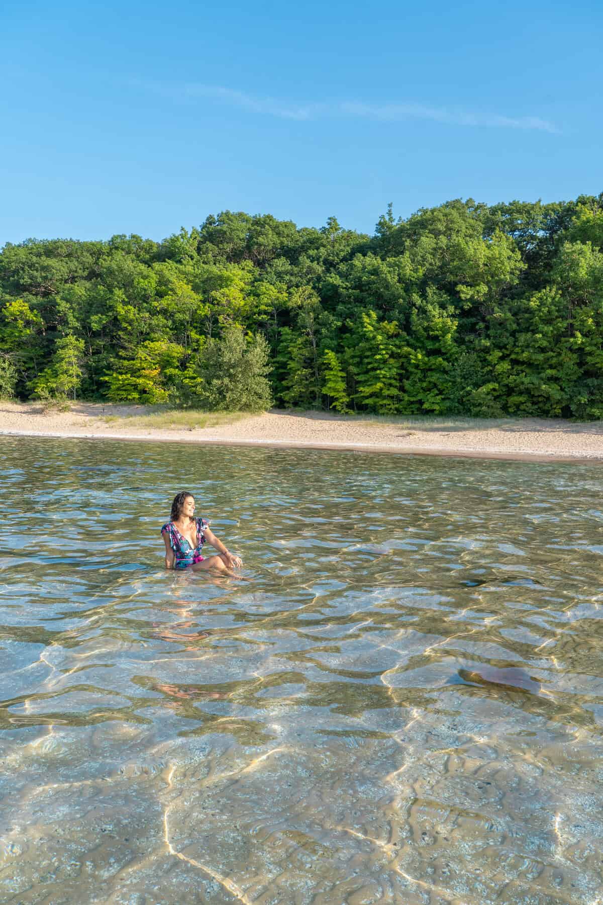 Person in water near a beach with a backdrop of trees under a clear sky.