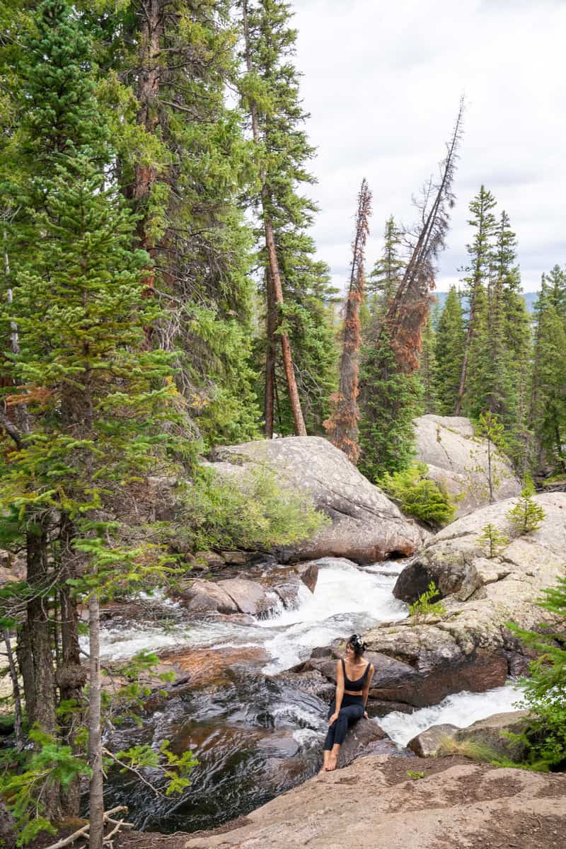 Person sitting on a rock by a forest stream with evergreen trees.