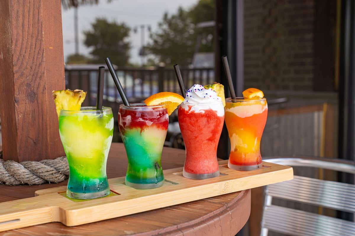 Four colorful layered cocktails on a wooden tray with garnishes.