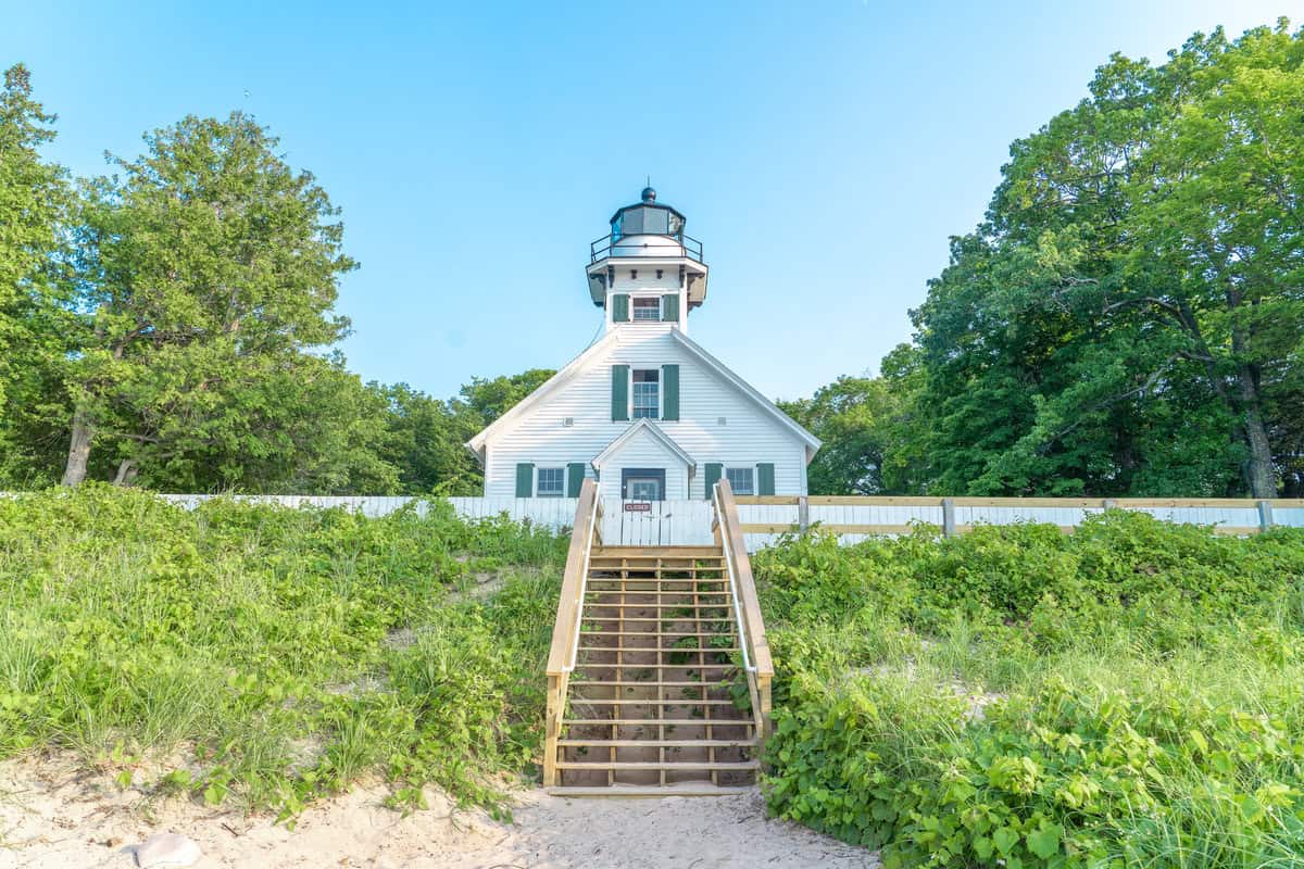 White lighthouse with a staircase surrounded by greenery under a clear blue sky.