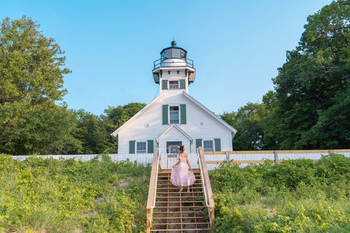 Person ascending stairs to a white lighthouse surrounded by greenery under a blue sky.