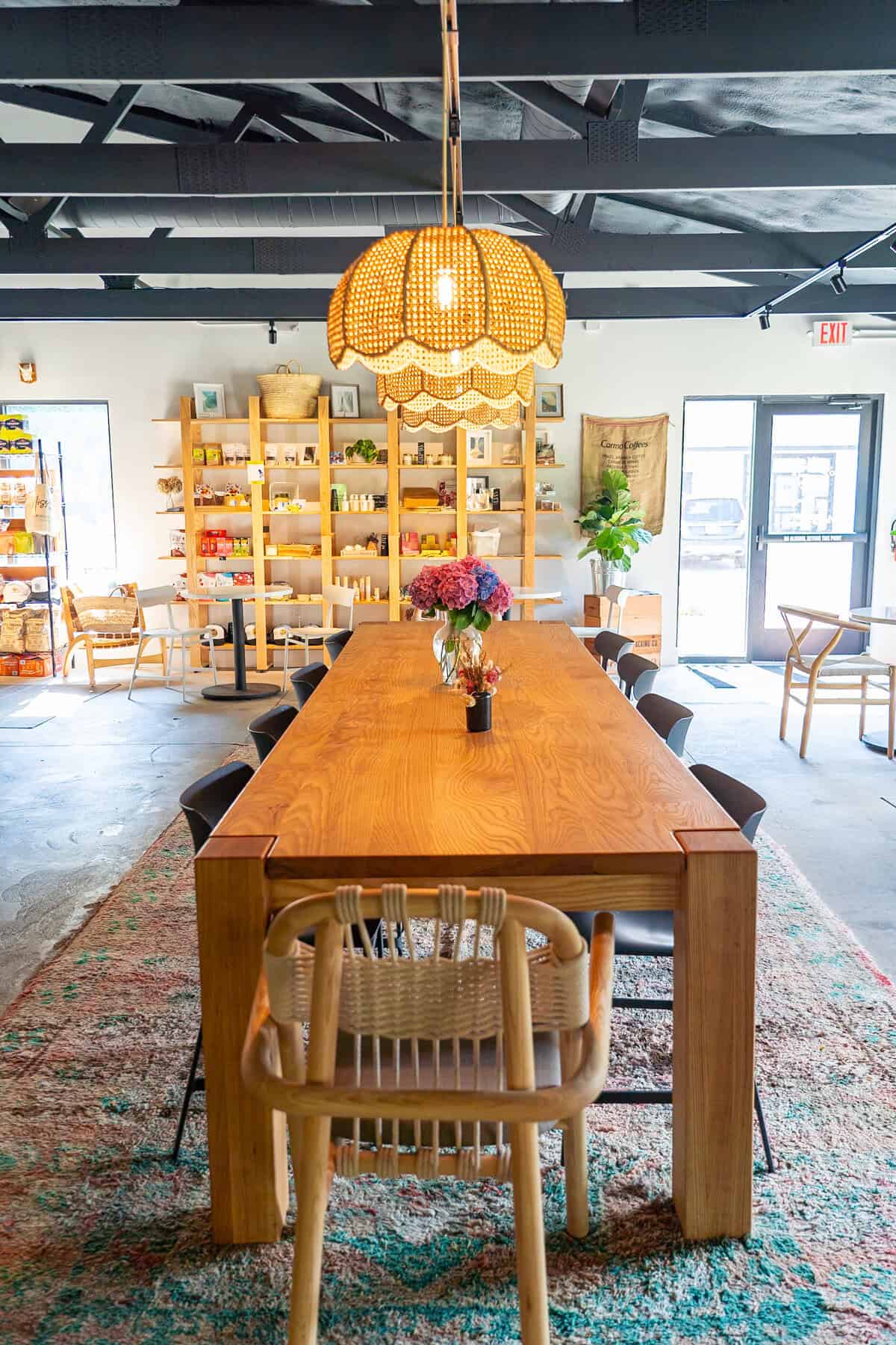 An inviting interior with a wooden table, chairs, wicker lamp, and shelf with products.