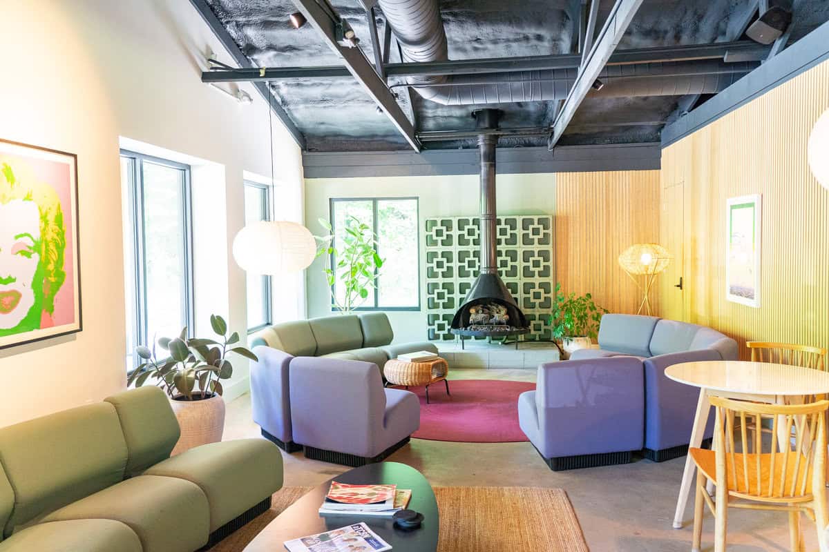 Modern living room with colorful furniture and a central fireplace, bright artwork on walls, and exposed ceiling.