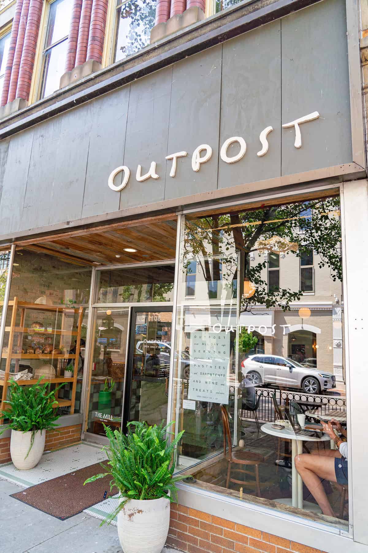 Storefront of "OUTPOST" with large windows, plants outside, and a person seated inside.