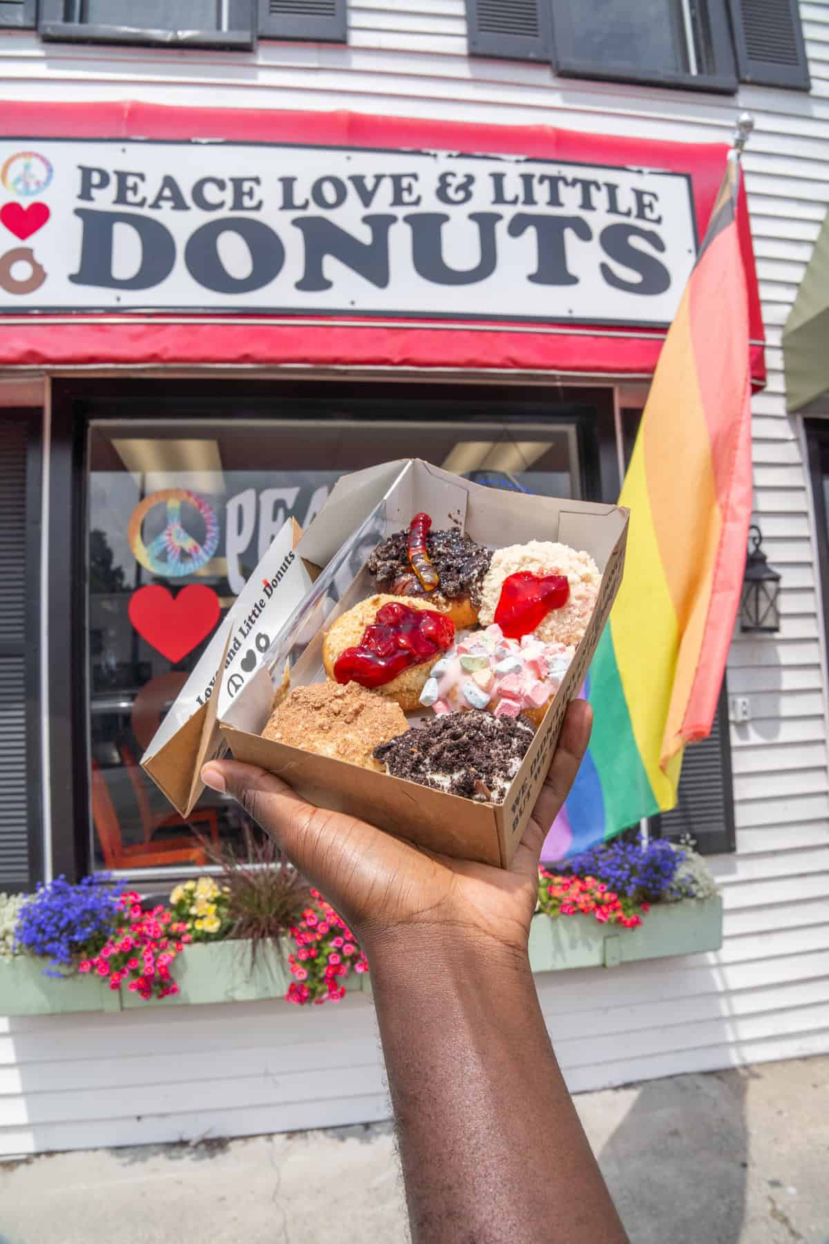 Hand holding a box of assorted donuts in front of a donut shop with a pride flag.