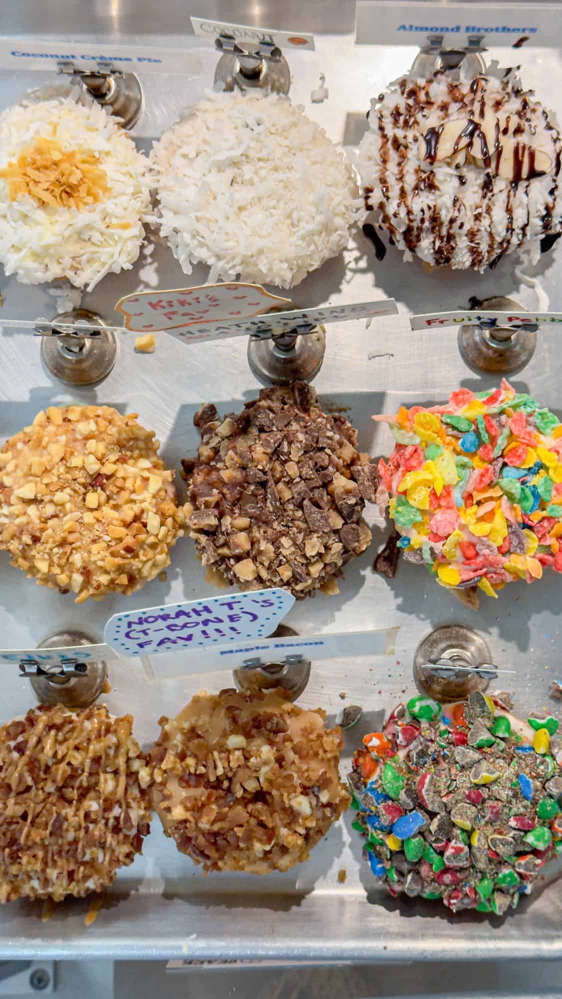 Assorted candy apples displayed on a shelf, with toppings like nuts, coconut, and colorful cereals.