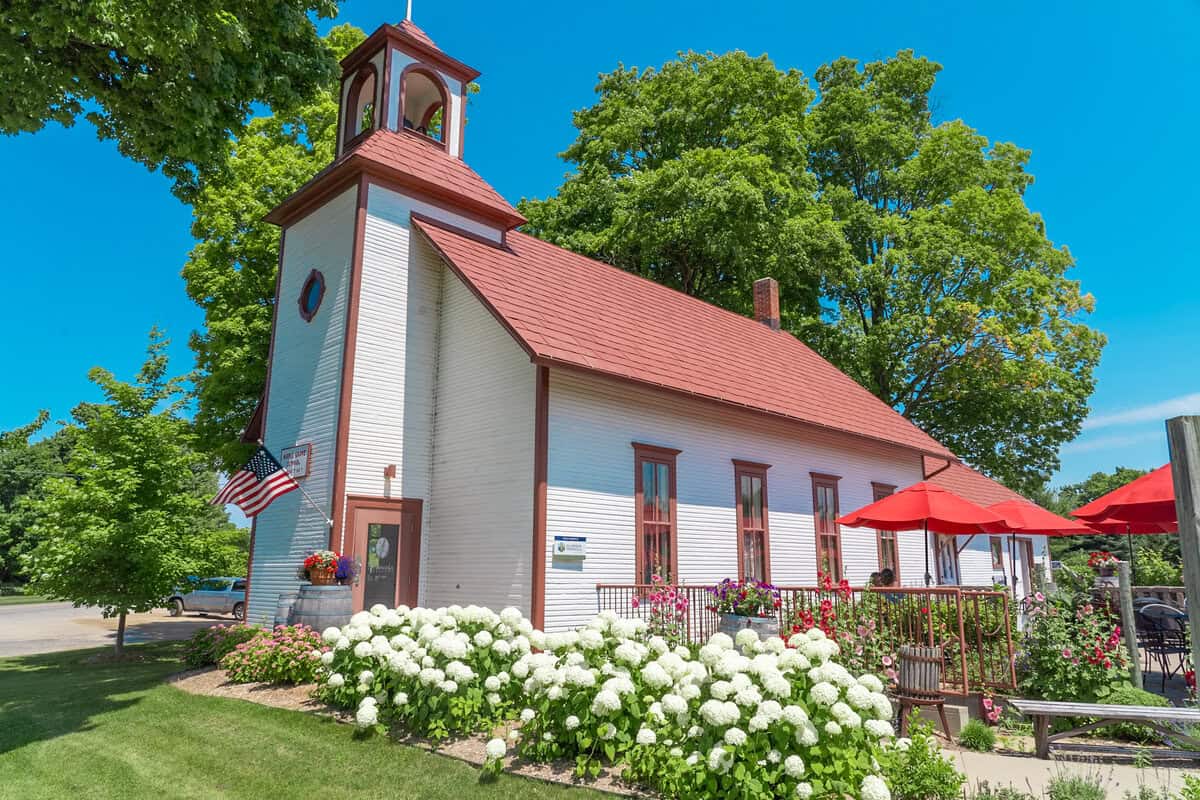 Quaint white church with red roof, bell tower, American flag, and surrounding flowers