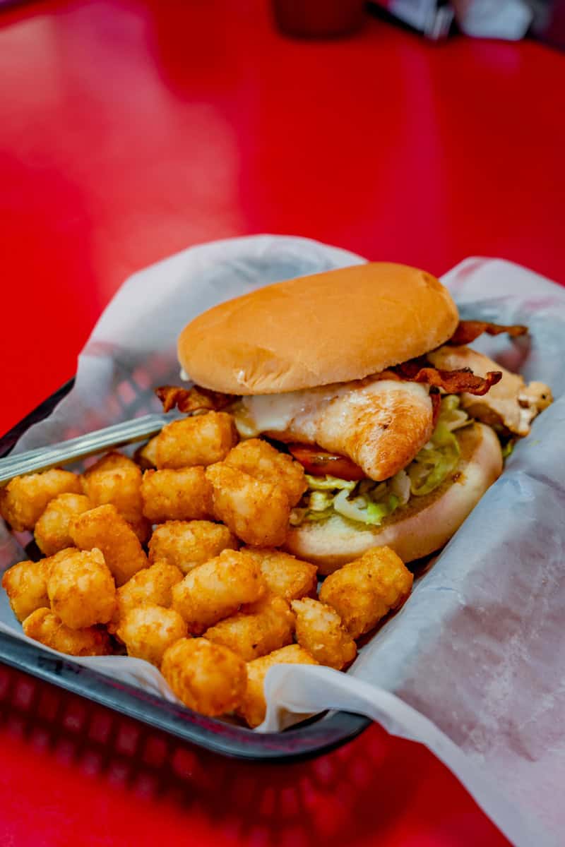 A chicken sandwich with bacon and tater tots on a red table.