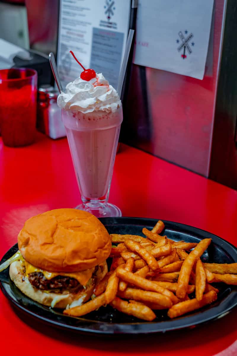 A cheeseburger with fries and a milkshake topped with whipped cream and a cherry, on a red table.