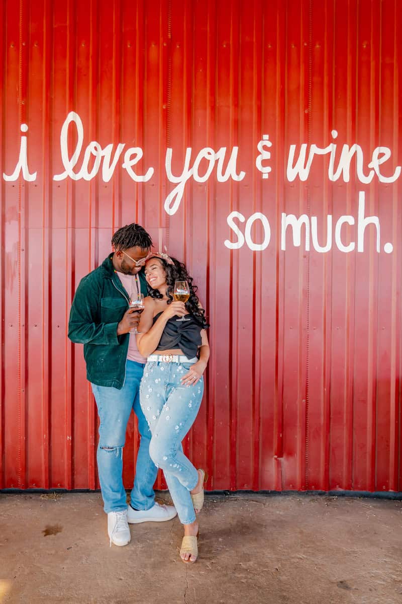 Couple embracing with wine glasses against a red wall with romantic text.