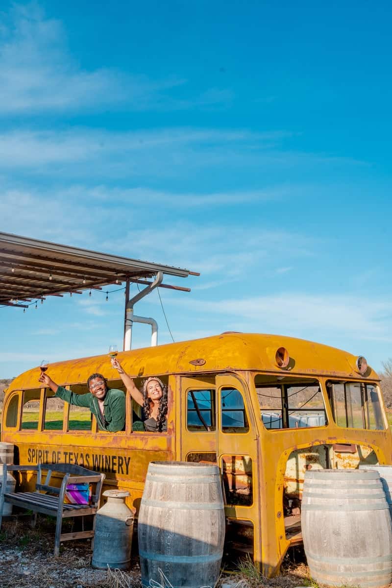 Yellow vintage bus converted into a bar at "Spirit of Texas Winery" with people and wine barrels.