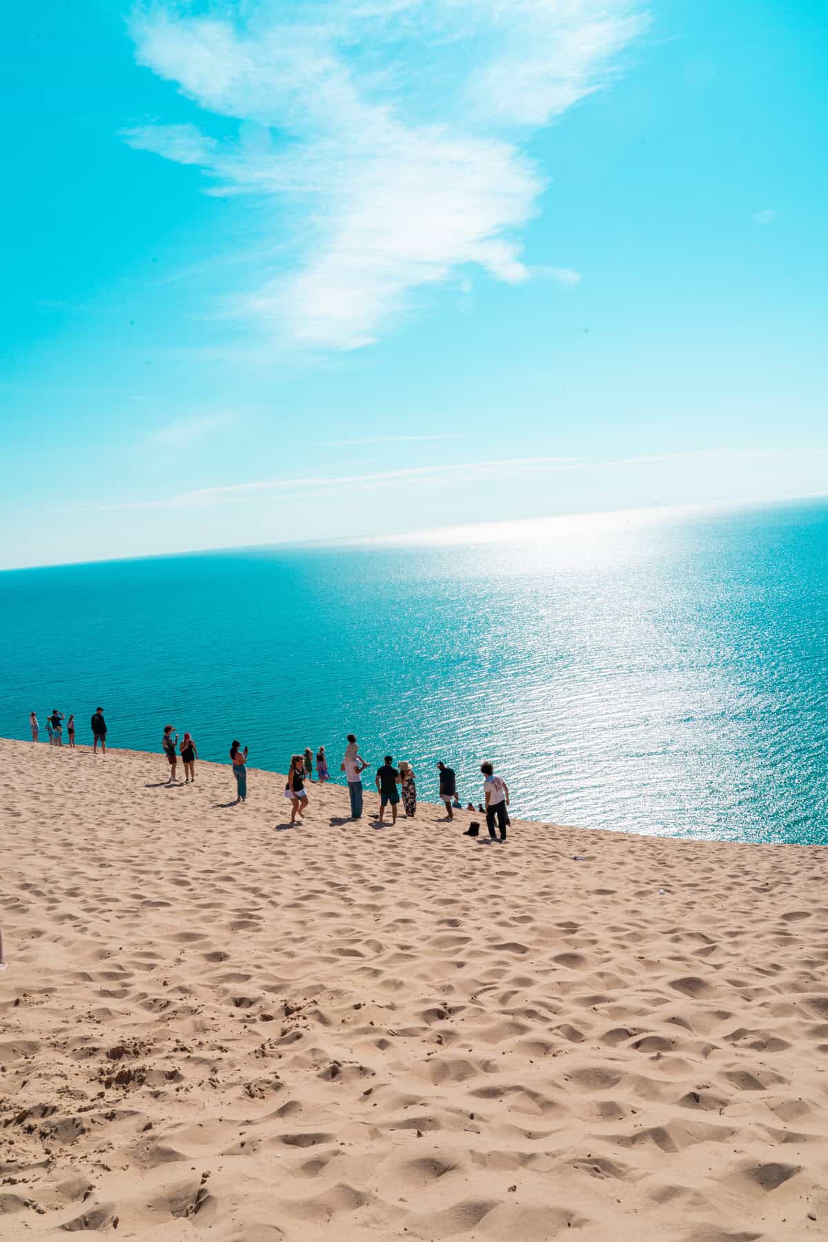 People on a sandy dune overlooking a sparkling blue sea under a sunny sky.