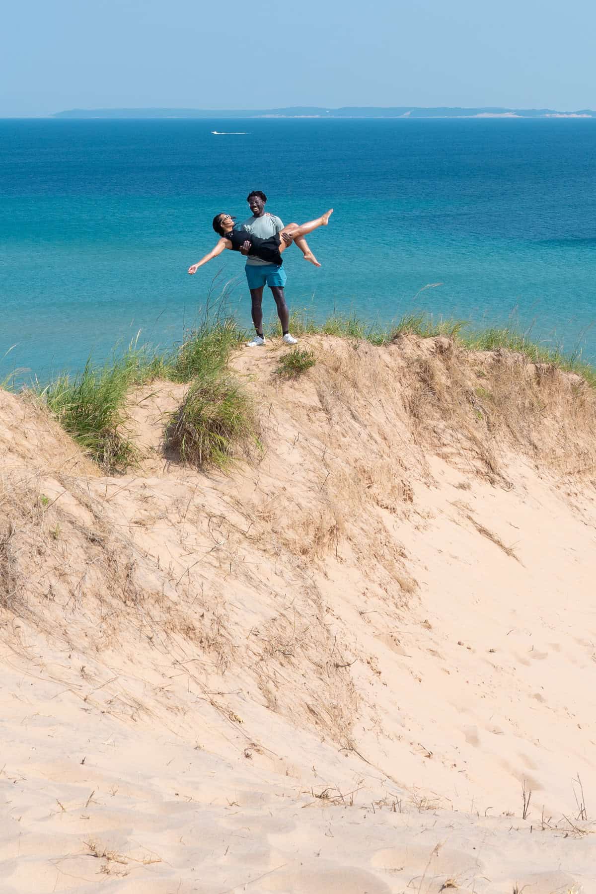 Person holding another in their arms on a sandy hill with the sea in the background.