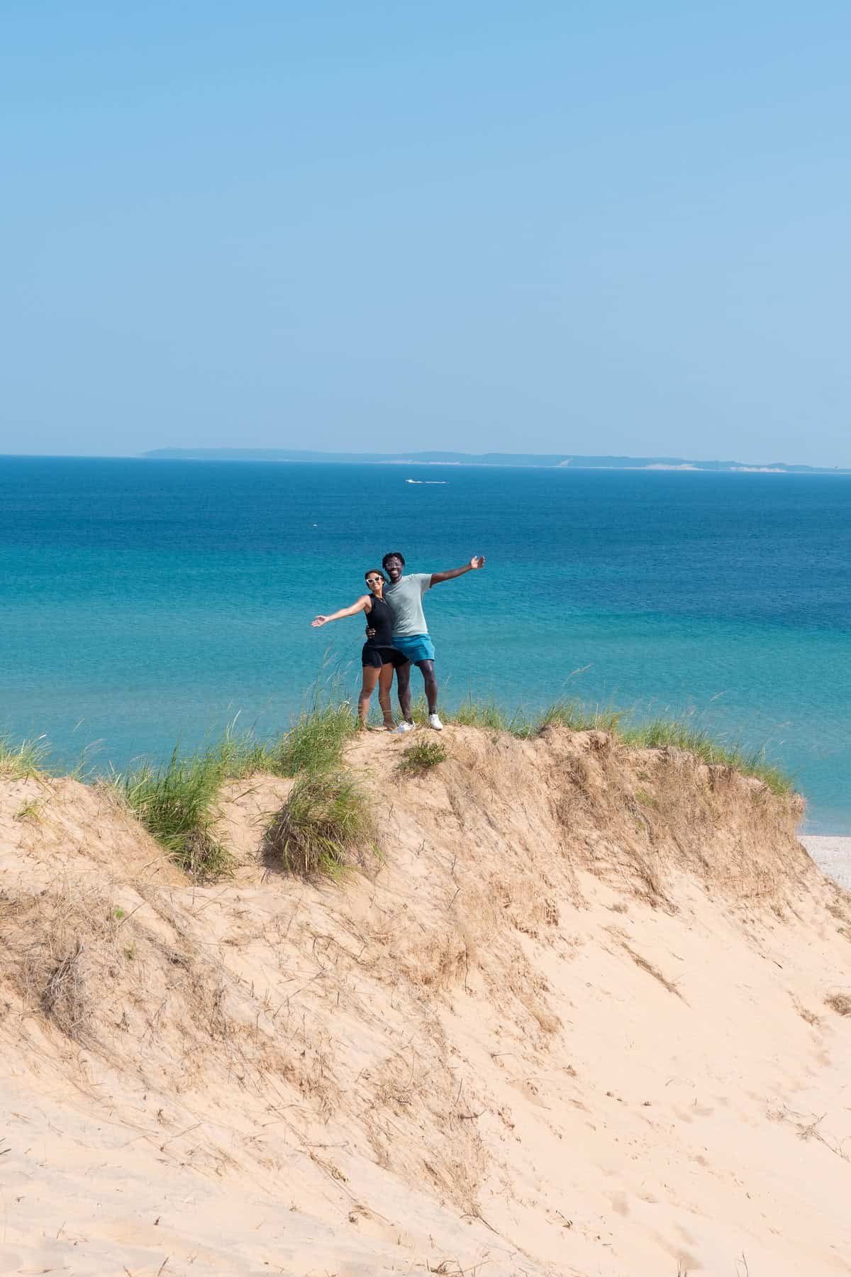 Two people standing on a sand dune overlooking a clear blue sea.