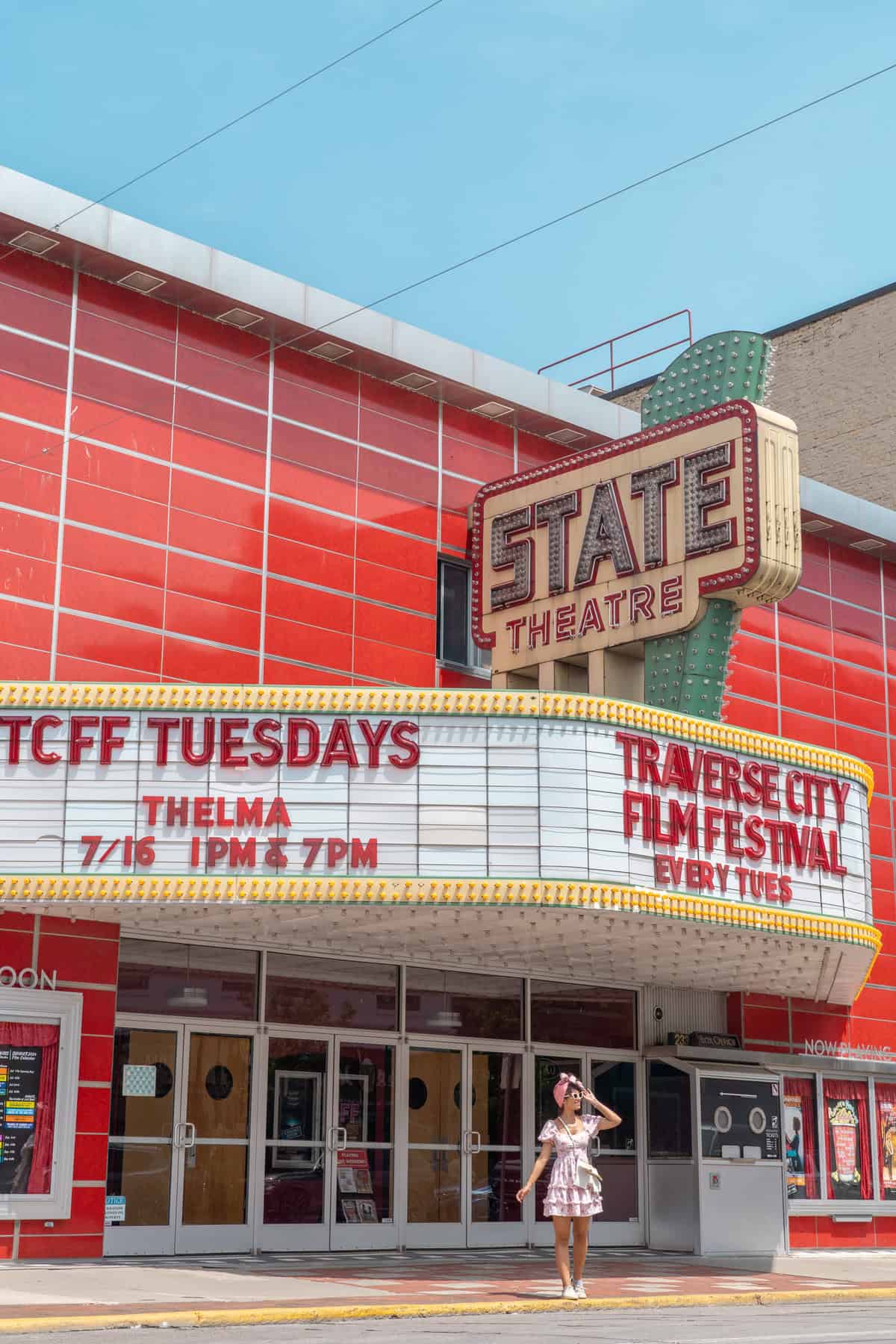 A person in a dress and hat standing in front of the State Theatre marquee on a sunny day.