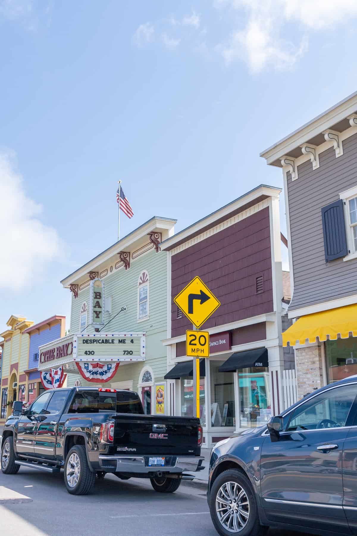 Colorful town street with a cinema marquee, American flag, a right-turn sign, and parked cars.