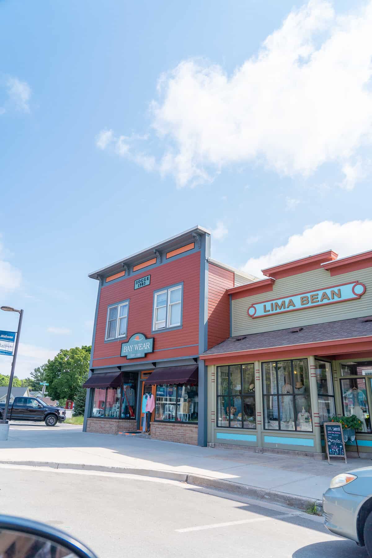 Colorful storefronts under a blue sky with light clouds, including a shop named "Bay Wear".