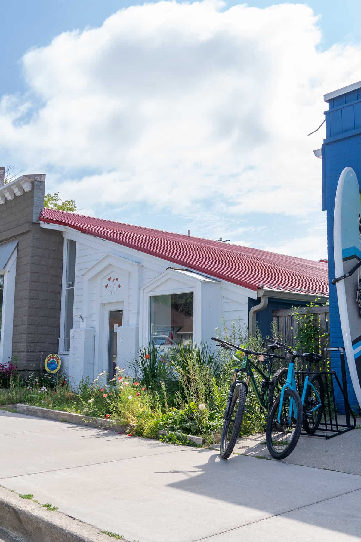 Small white buildings with a red roof, bicycles, and a surfboard against a blue sky.