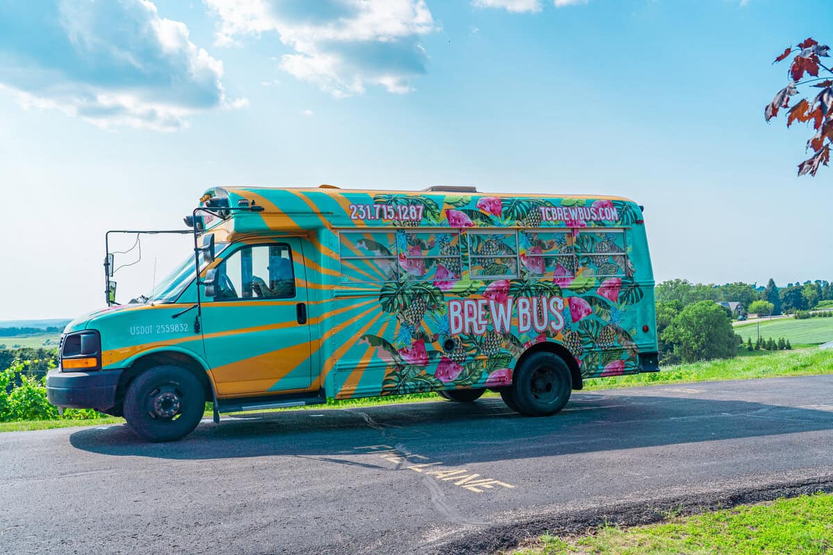 Colorful bus with 'BREW BUS' design parked on a road near a green field under a blue sky.