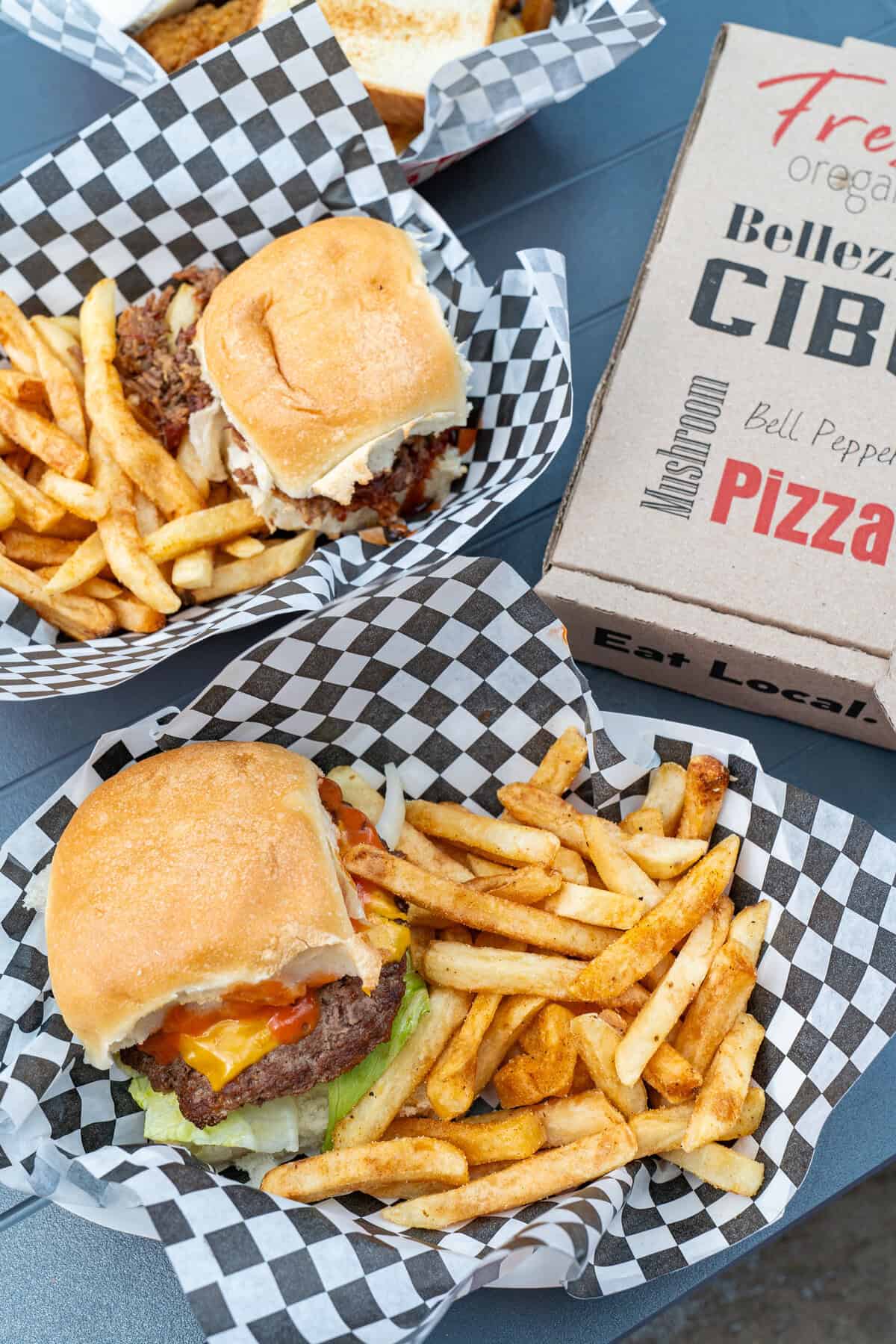 Two cheeseburgers with fries and a pizza box on a table.