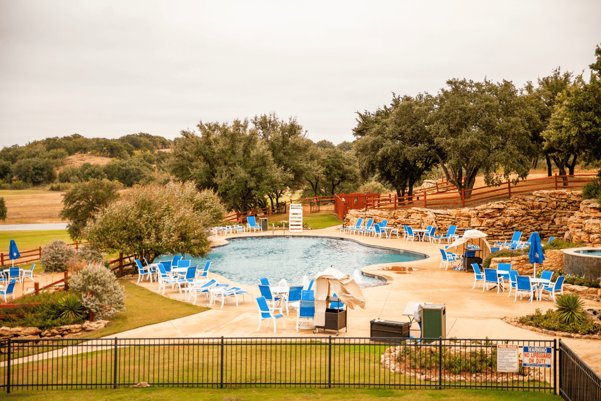 Outdoor swimming pool with blue loungers surrounded by trees and a fenced lawn area.