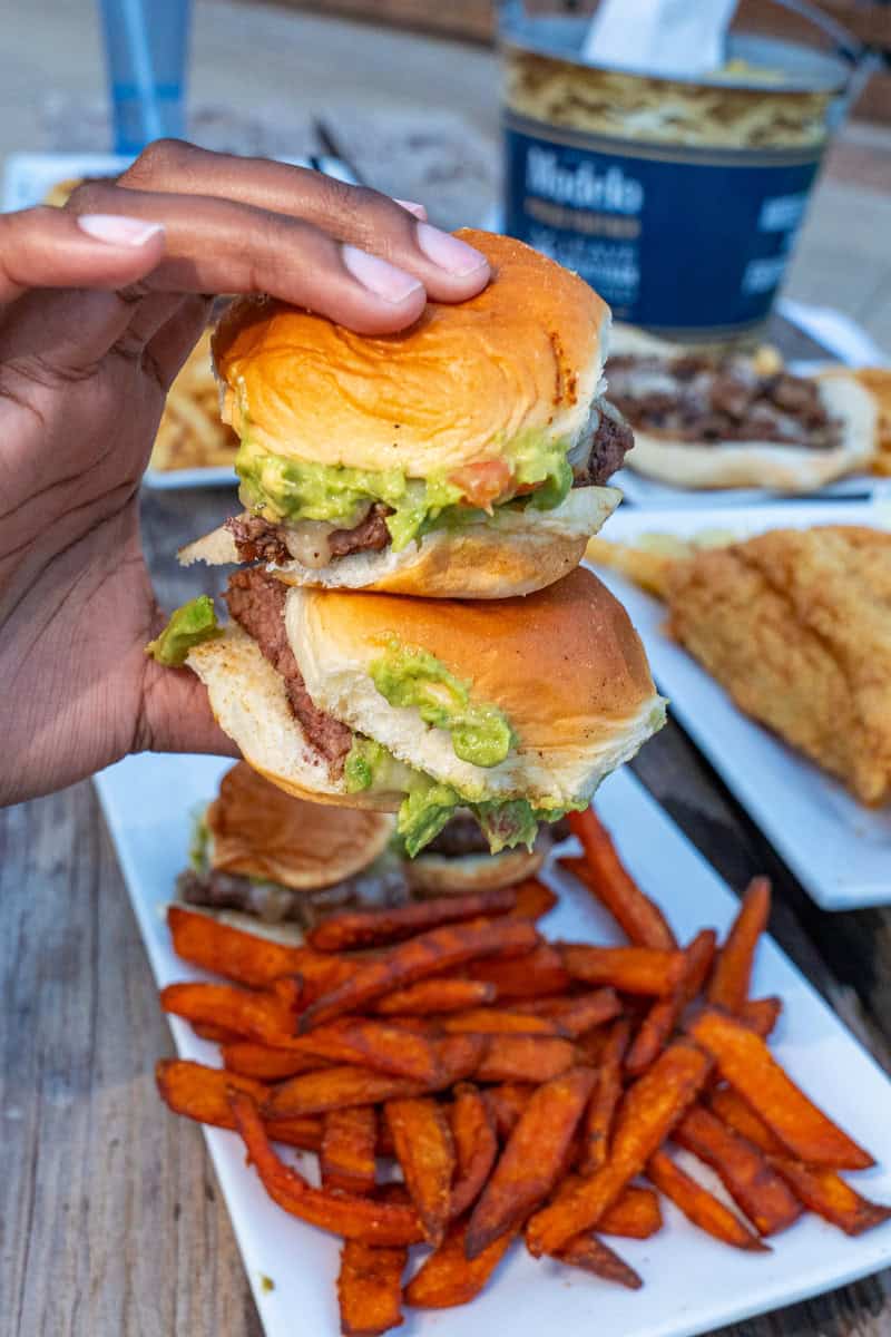 A hand holding a bitten burger over a plate of sweet potato fries.